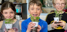 Three early years students facing camera and smiling, each holding a potted plant from their Little Green Thumbs garden